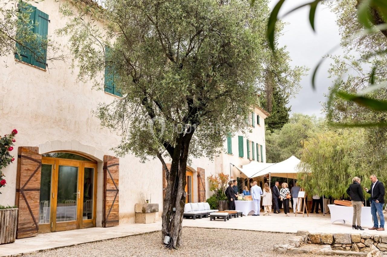 Un groupe de personnes discutant près d'une maison en pierre avec volets en bois, entourée d'arbres et de végétation.