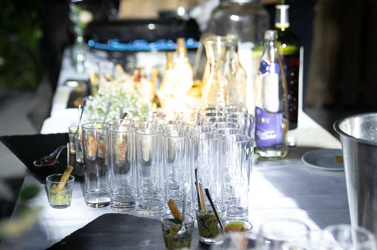 Table de service en extérieur avec verres vides alignés, amuse-bouches et un chef cuisinant en arrière-plan.
