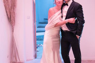 Un couple habillé élégamment, la femme en robe blanche et l'homme en costume, pose dans une pièce lumineuse.