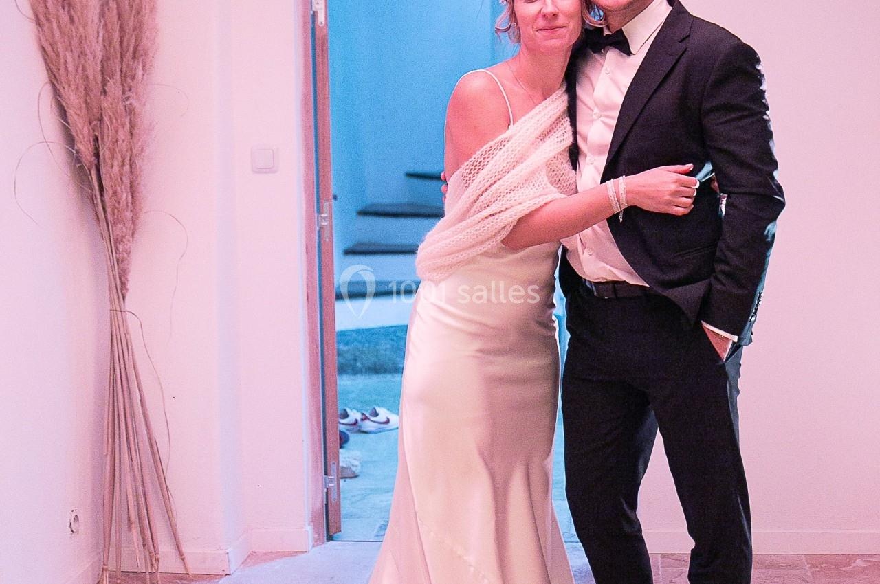 Un couple habillé élégamment, la femme en robe blanche et l'homme en costume, pose dans une pièce lumineuse.