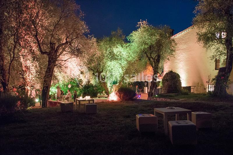 Jardin éclairé de nuit avec des arbres illuminés, des tables basses et des poufs disposés sur une pelouse.