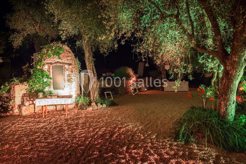Cour éclairée de nuit avec des arbres, un mur en pierre, des fleurs rouges et un banc sous un éclairage chaleureux.