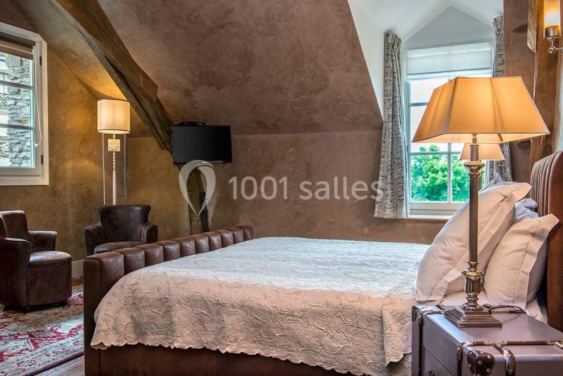 Chambre mansardée avec lit double, fauteuils en cuir, lampes allumées et fenêtre donnant sur un paysage verdoyant.