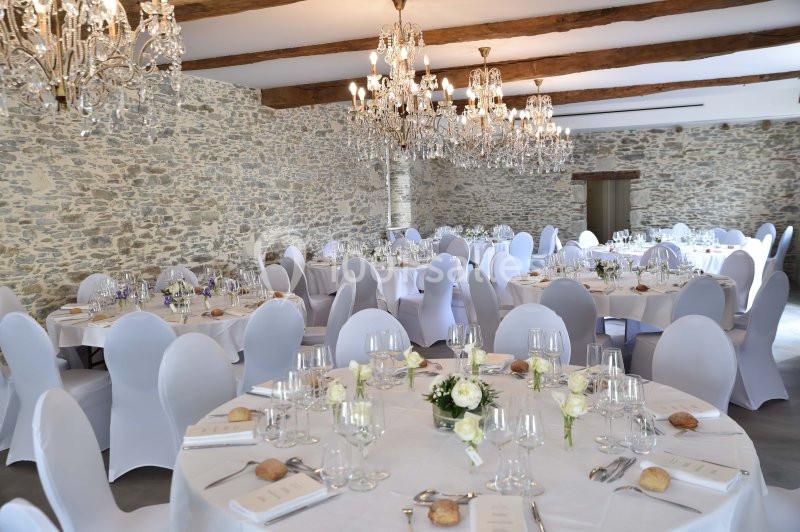 Salle de réception avec murs en pierre, tables rondes dressées, nappes blanches et lustres en cristal suspendus.