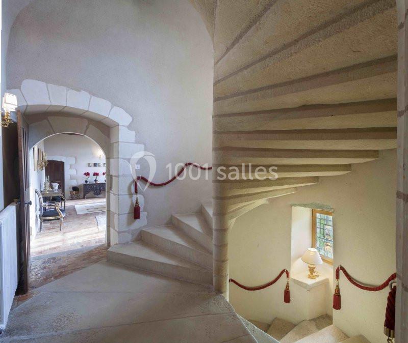 Escalier en pierre avec rampe ornée de cordons rouges, menant à une pièce lumineuse avec fenêtre et lampe.