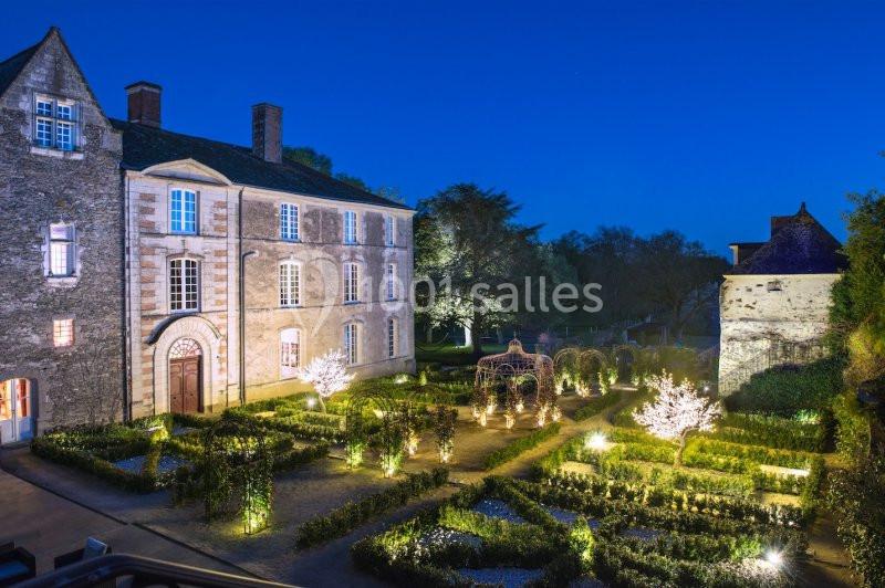 Jardin éclairé avec des arbustes taillés et des arbres illuminés, devant un bâtiment historique en soirée.