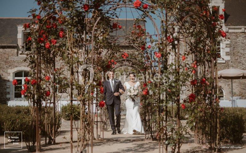 Un couple marche sous une arche fleurie de roses rouges dans un jardin devant un bâtiment en pierre.