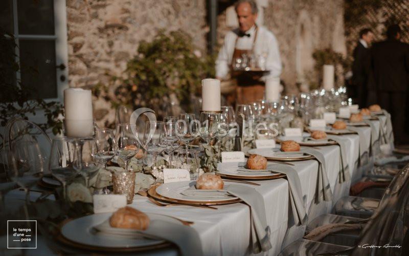Table élégamment dressée en extérieur avec nappes blanches, vaisselle raffinée, bougies et petits pains individuels.