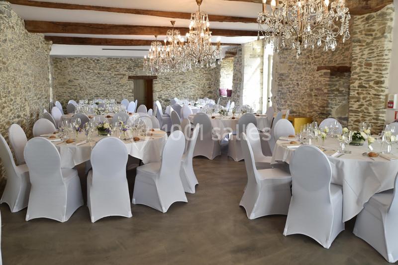 Salle de réception avec murs en pierre, tables rondes dressées et chaises blanches sous des lustres élégants.