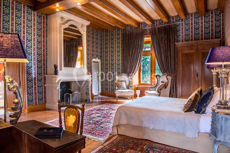 Chambre élégante avec lit double, cheminée en pierre, fauteuils anciens, tapis coloré et grandes fenêtres avec rideaux.