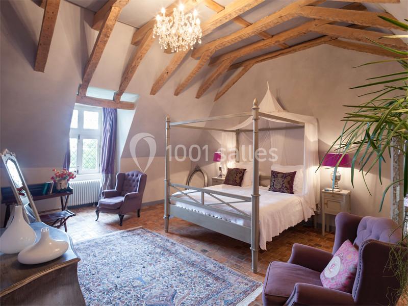 Chambre lumineuse avec lit à baldaquin, poutres apparentes, fauteuils violets et décoration élégante.