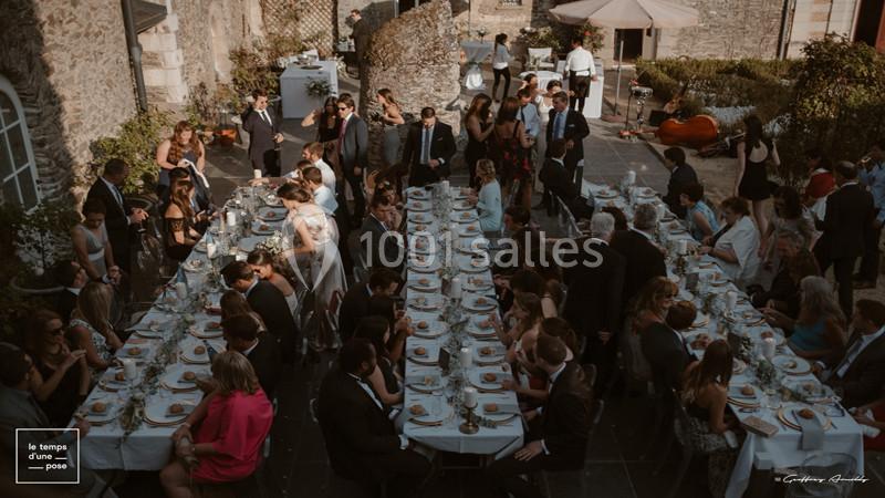 Invités assis à de longues tables dressées en extérieur lors d'une réception élégante dans une cour en pierre.