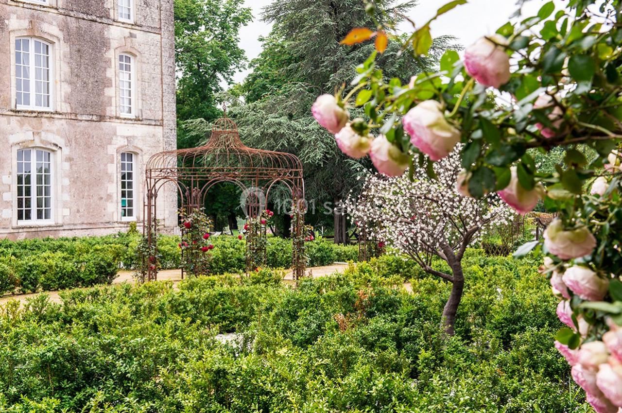 Jardin fleuri avec une structure métallique ornée de roses, entourée de buissons et d'arbres en arrière-plan.
