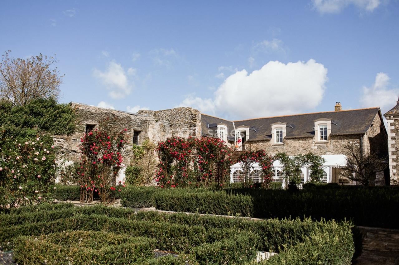 Façade en pierre d'une bâtisse ancienne entourée de rosiers grimpants et d'un jardin structuré.