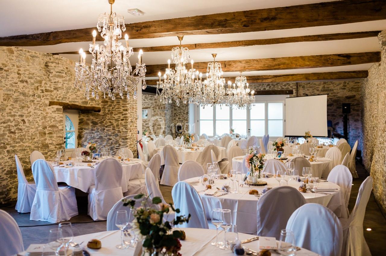 Salle de réception avec murs en pierre, tables rondes dressées, chaises blanches et lustres suspendus.