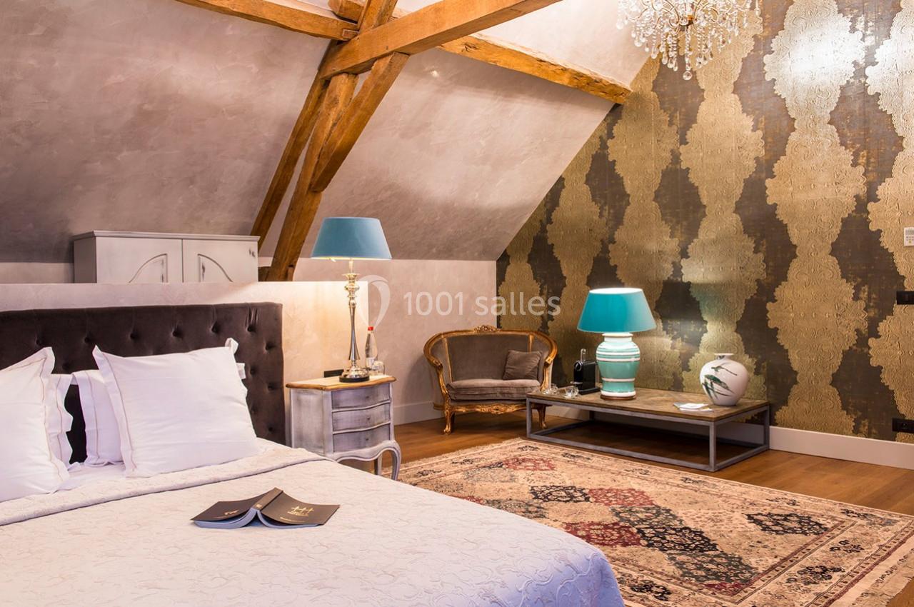 Chambre mansardée avec lit double, poutres apparentes, tapis, lampes bleues et fauteuil en bois dans un décor élégant.