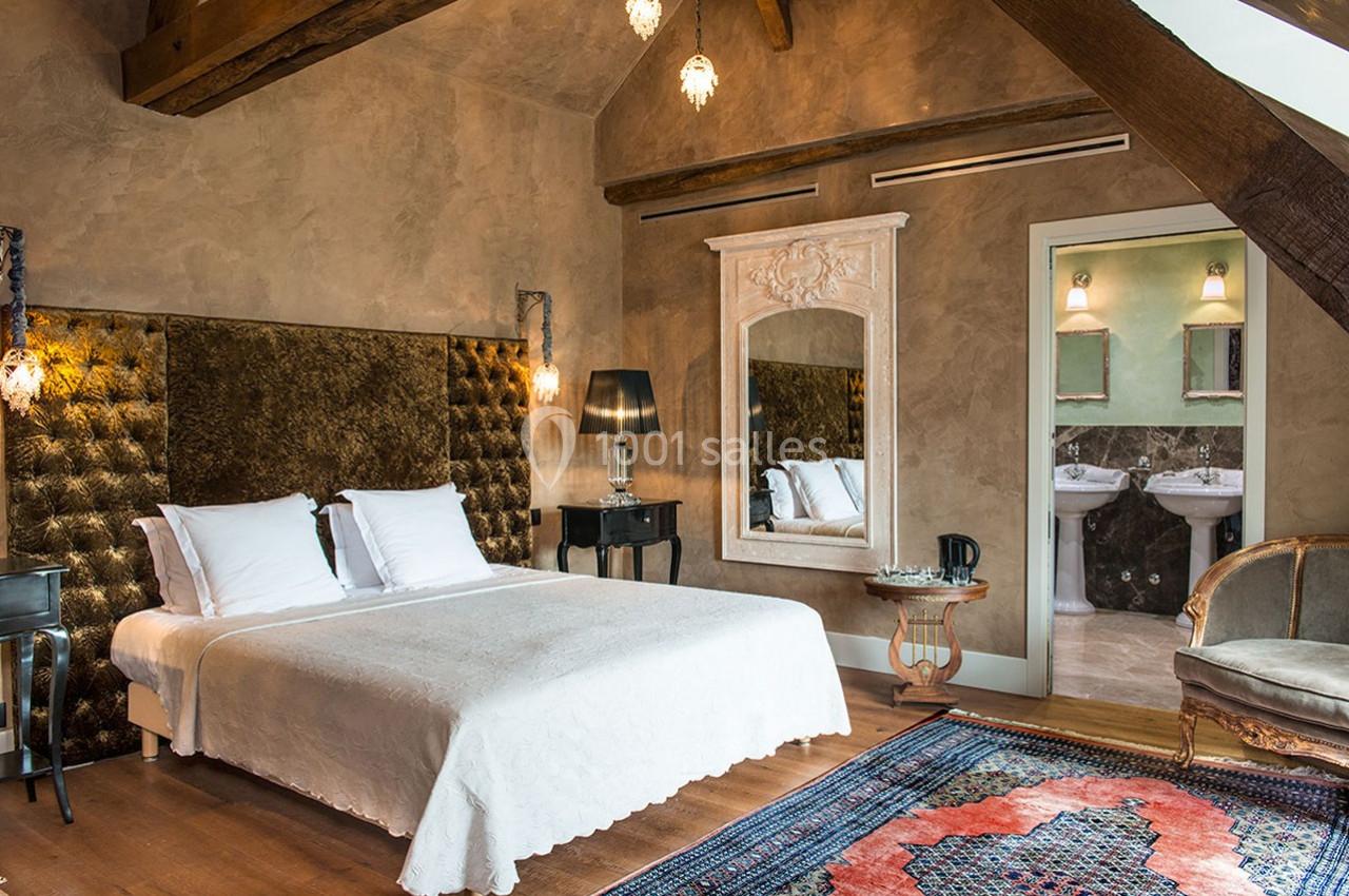 Chambre lumineuse avec lit double, murs texturés, miroir décoratif, poutres apparentes et vue sur une salle de bain…