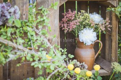 Bouquet de fleurs blanches et roses dans un pichet en terre cuite, posé dans une caisse en bois entourée de verdure.