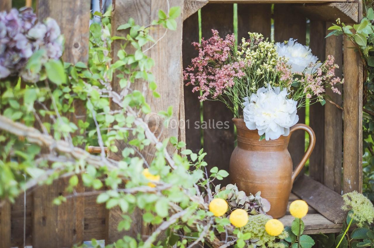 Bouquet de fleurs blanches et roses dans un pichet en terre cuite, posé dans une caisse en bois entourée de verdure.