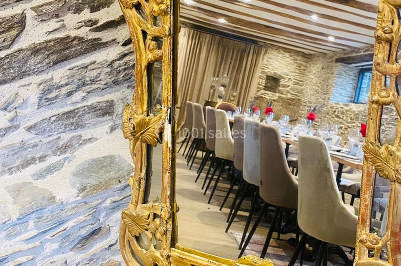 Reflet d'une salle à manger élégante avec table dressée, chaises beige et murs en pierre, dans un miroir doré sculpté.