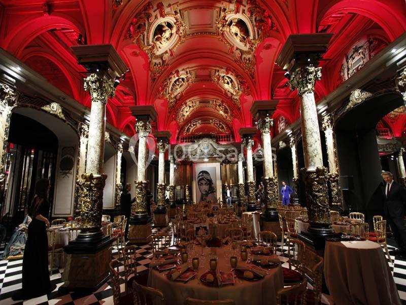 Salle de réception luxueuse avec colonnes dorées, plafond rouge orné et tables dressées sur un sol en damier.