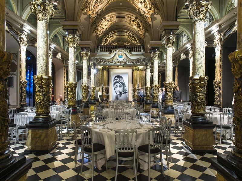 Salle ornée de colonnes dorées et de fresques, aménagée avec des tables rondes et des chaises pour un événement.