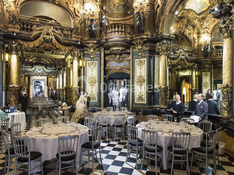 Salle de réception luxueuse avec tables rondes dressées, décor baroque doré et sol en damier noir et blanc.