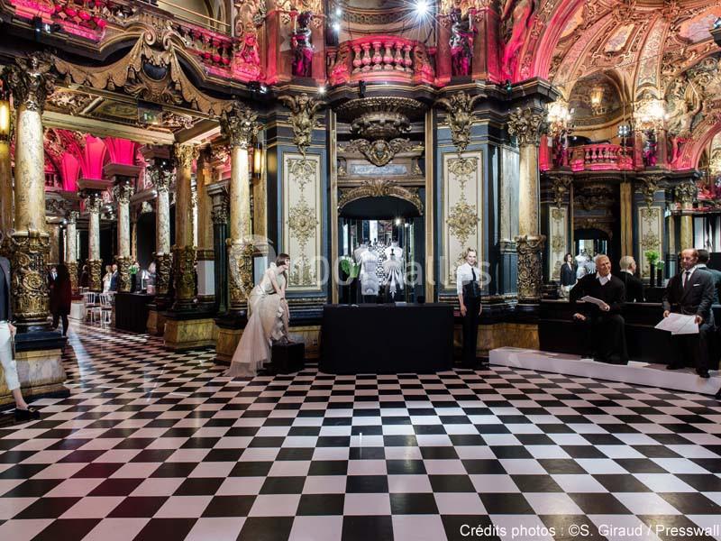 Salle luxueuse avec sol en damier, colonnes dorées, décorations baroques et éclairage rose, avec quelques personnes…