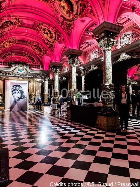 Intérieur d'un bâtiment avec plafond voûté illuminé en rose, sol à carreaux noirs et blancs, et affiches artistiques.
