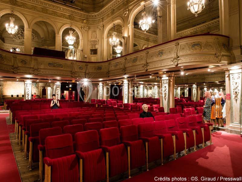 Salle de théâtre avec sièges rouges, balcons ornés et lustres suspendus, quelques personnes présentes.