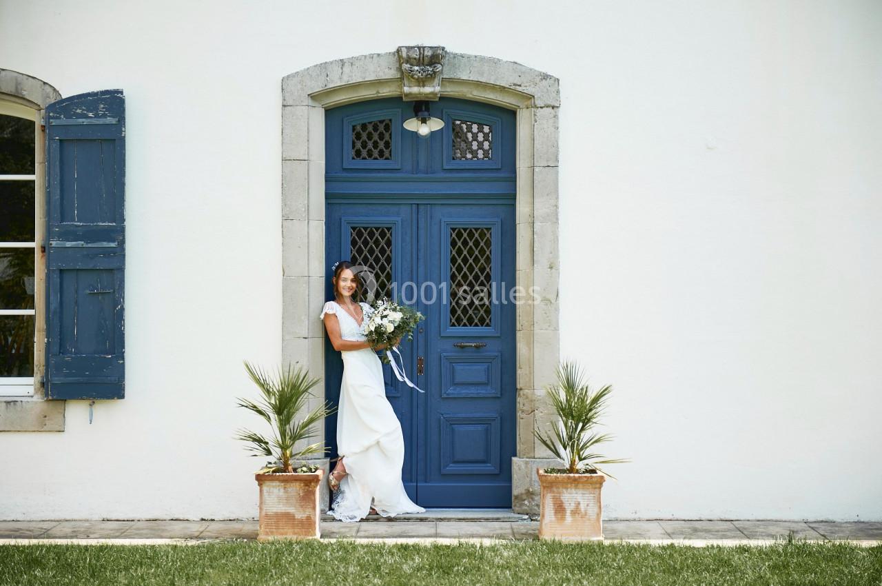 Femme en robe de mariée tenant un bouquet devant une porte bleue encadrée de volets et de plantes en pot.