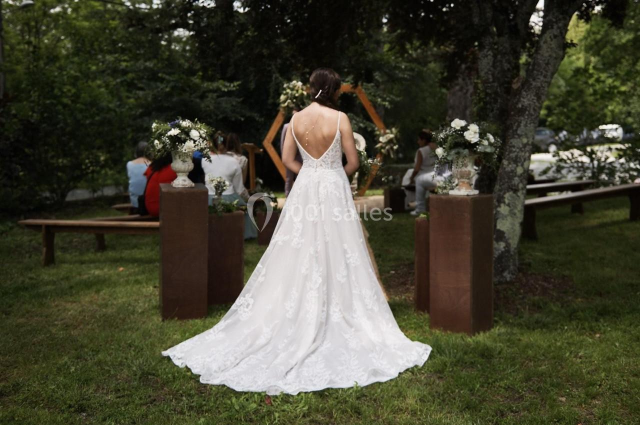 Une mariée en robe blanche vue de dos, debout dans un jardin devant une arche décorée pour une cérémonie en plein air.