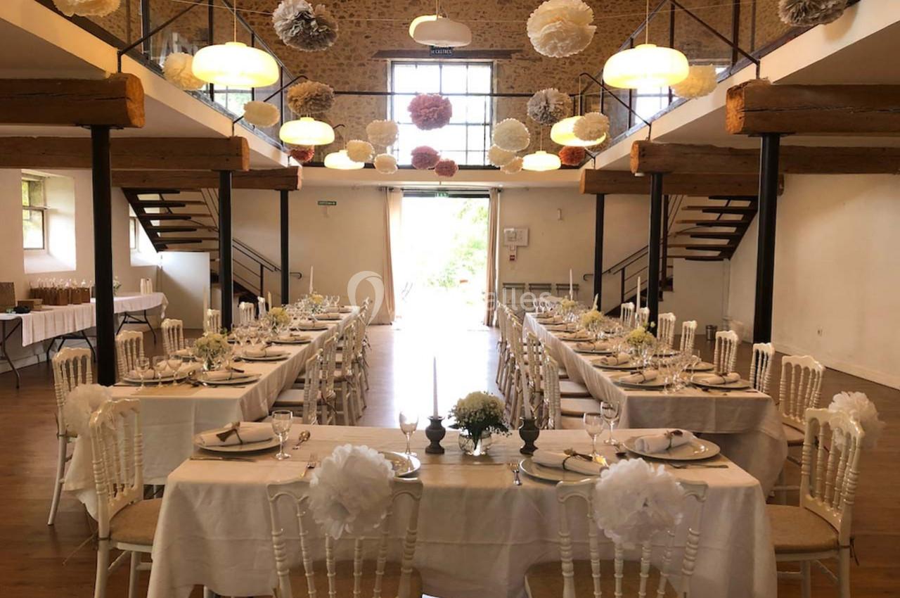 Salle de réception décorée pour un mariage, avec longues tables dressées, chaises blanches et suspensions en papier.