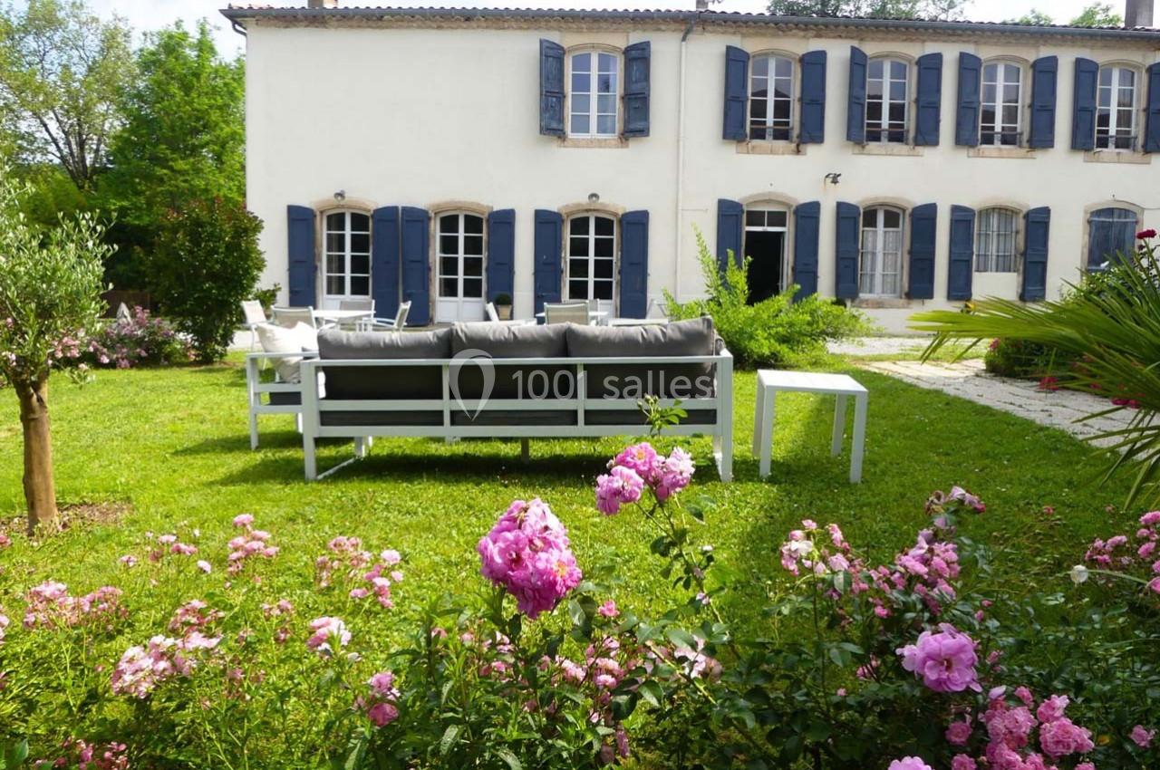 Façade d'une maison blanche avec volets bleus, jardin fleuri au premier plan et mobilier de jardin sur la pelouse.