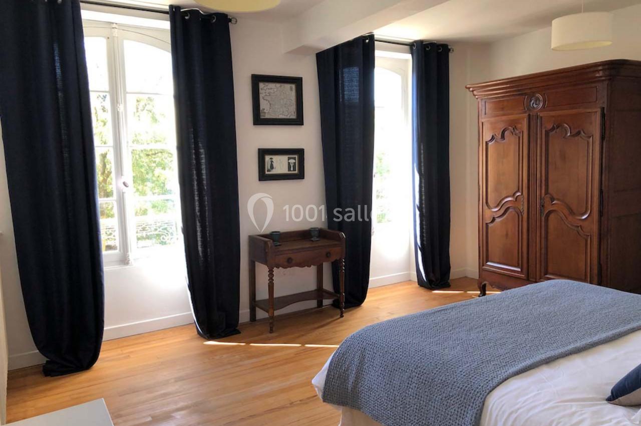 Chambre lumineuse avec parquet, armoire en bois, lit avec couverture bleue et rideaux sombres encadrant deux fenêtres.