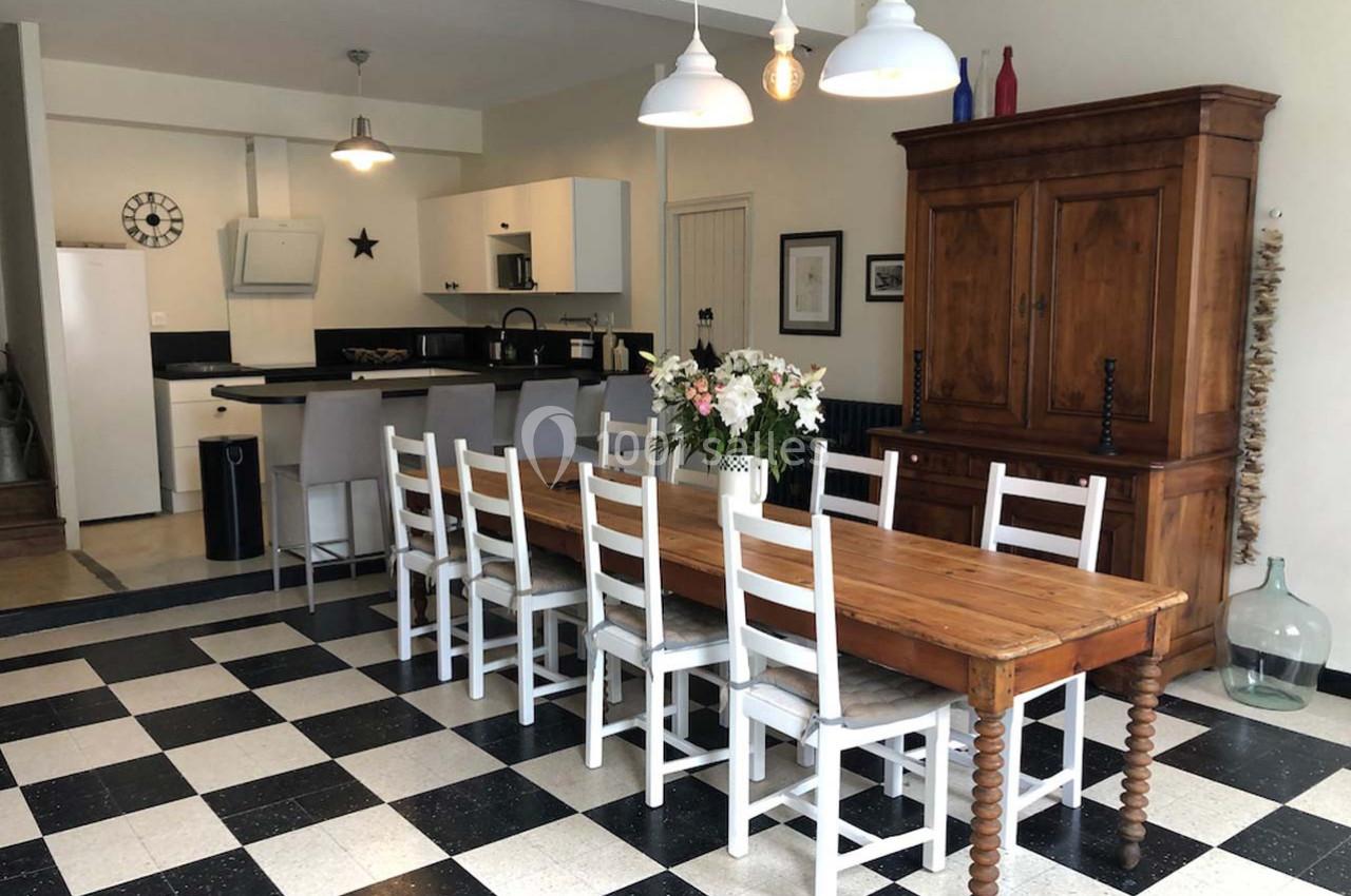 Salle à manger avec une grande table en bois, chaises blanches, cuisine ouverte et sol en damier noir et blanc.