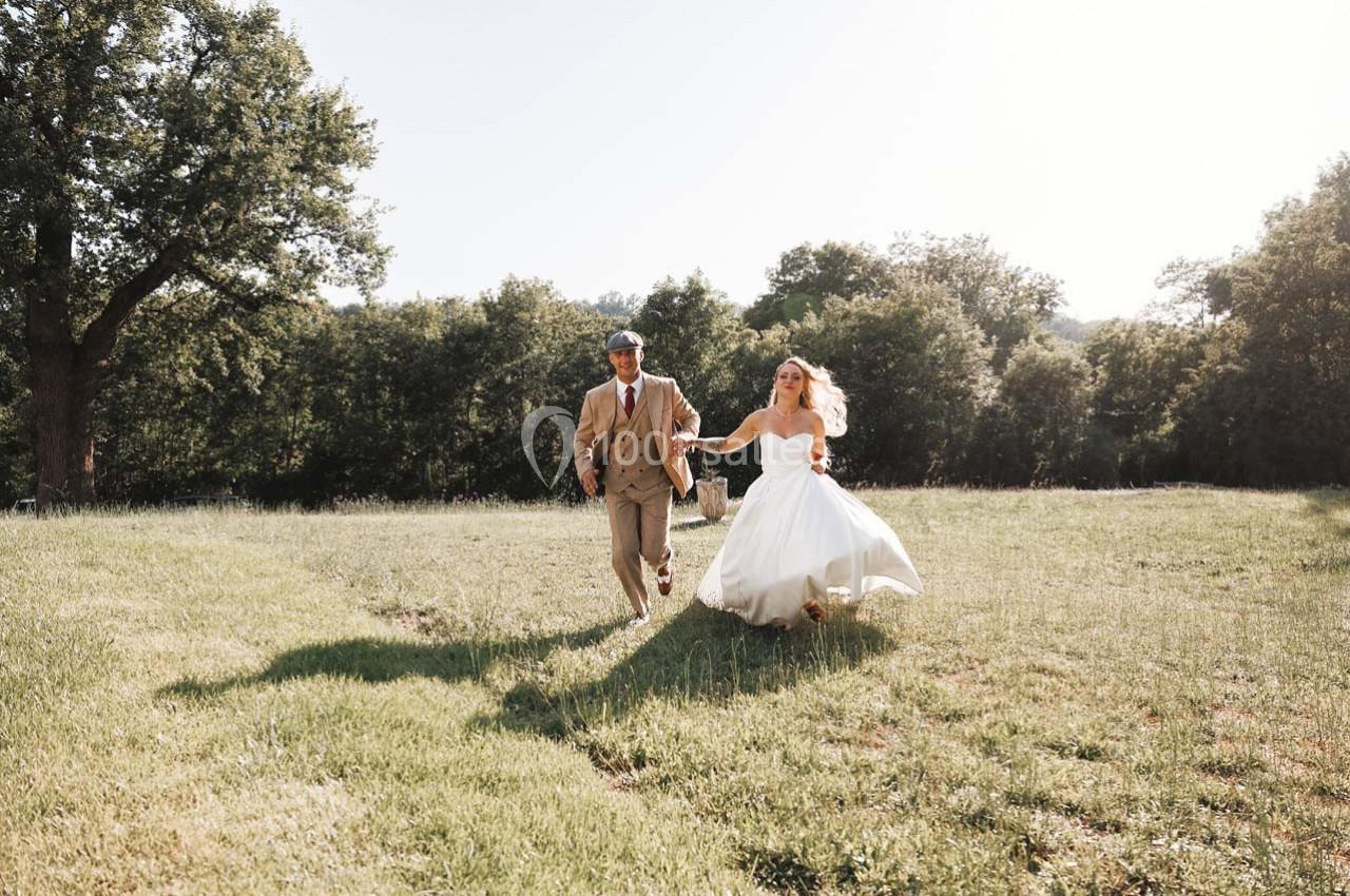 Un couple en tenue de mariage marche main dans la main dans un champ verdoyant sous une lumière naturelle.