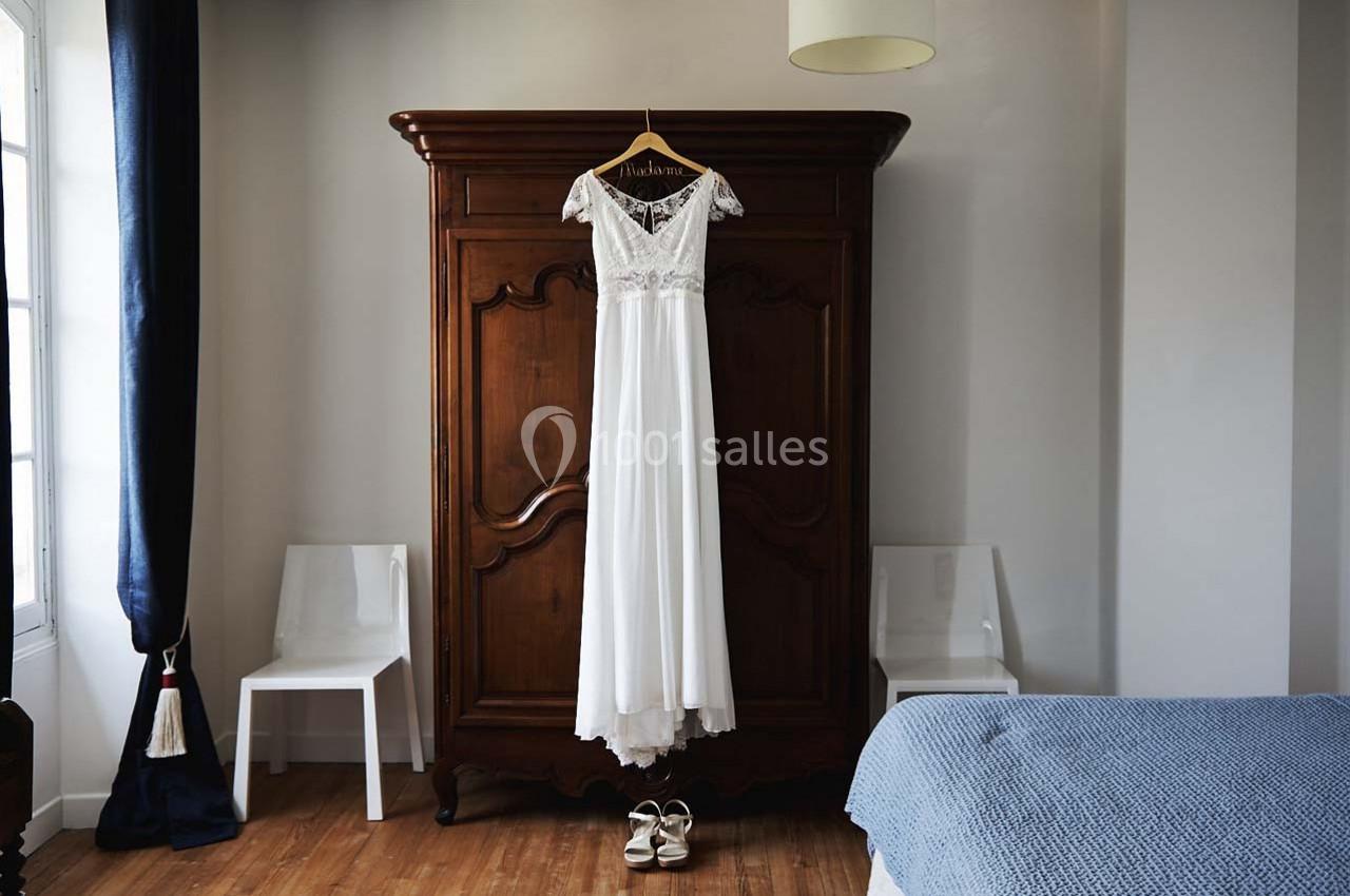 Robe de mariée blanche suspendue à une armoire en bois dans une chambre lumineuse avec parquet.