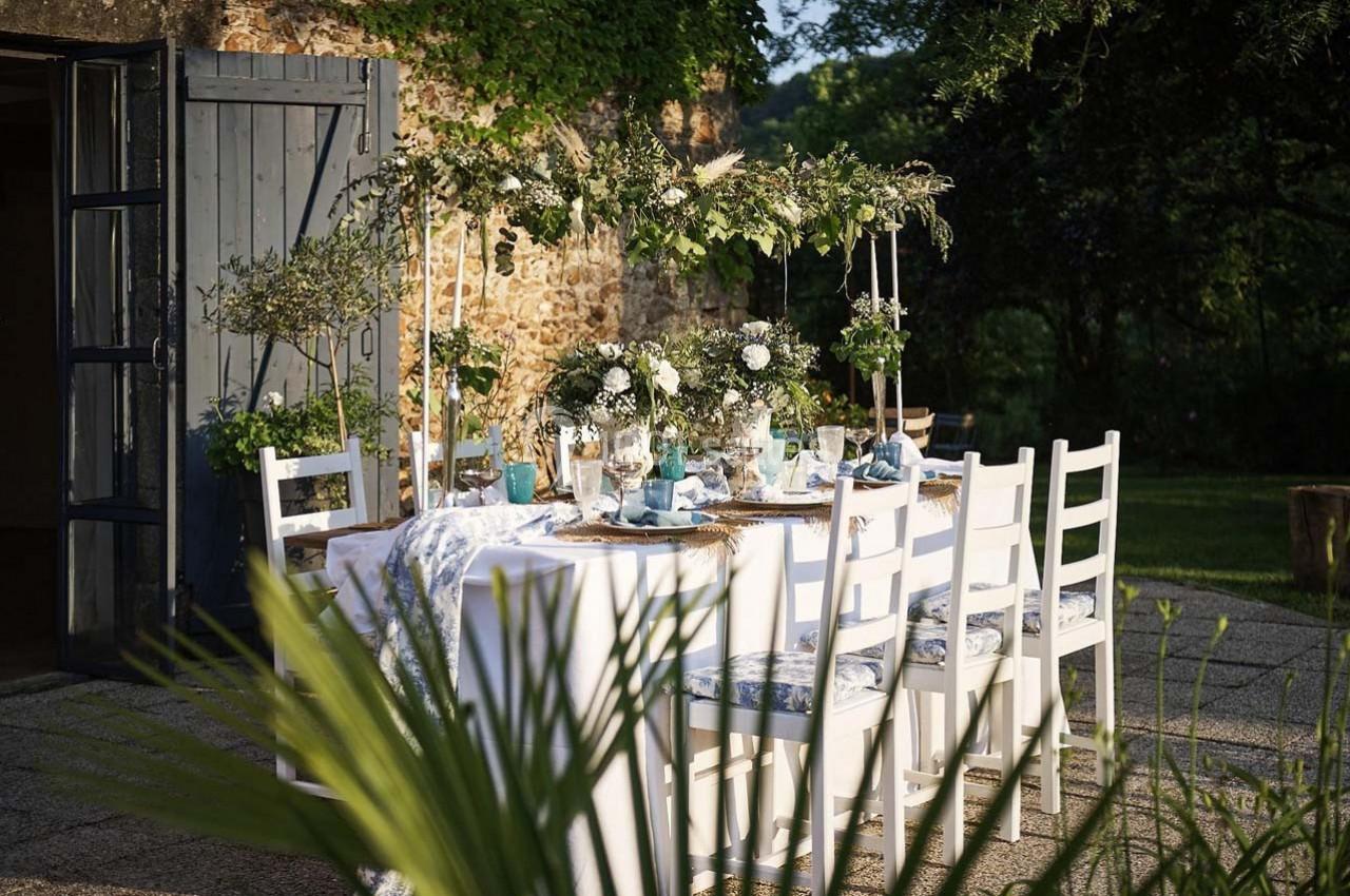 Table extérieure décorée avec des fleurs blanches, vaisselle élégante et chaises blanches, dans un jardin ensoleillé.