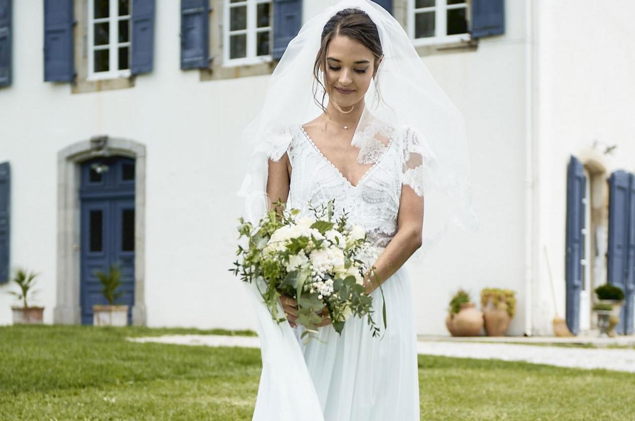 Une mariée en robe blanche tenant un bouquet de fleurs devant un bâtiment aux volets bleus.