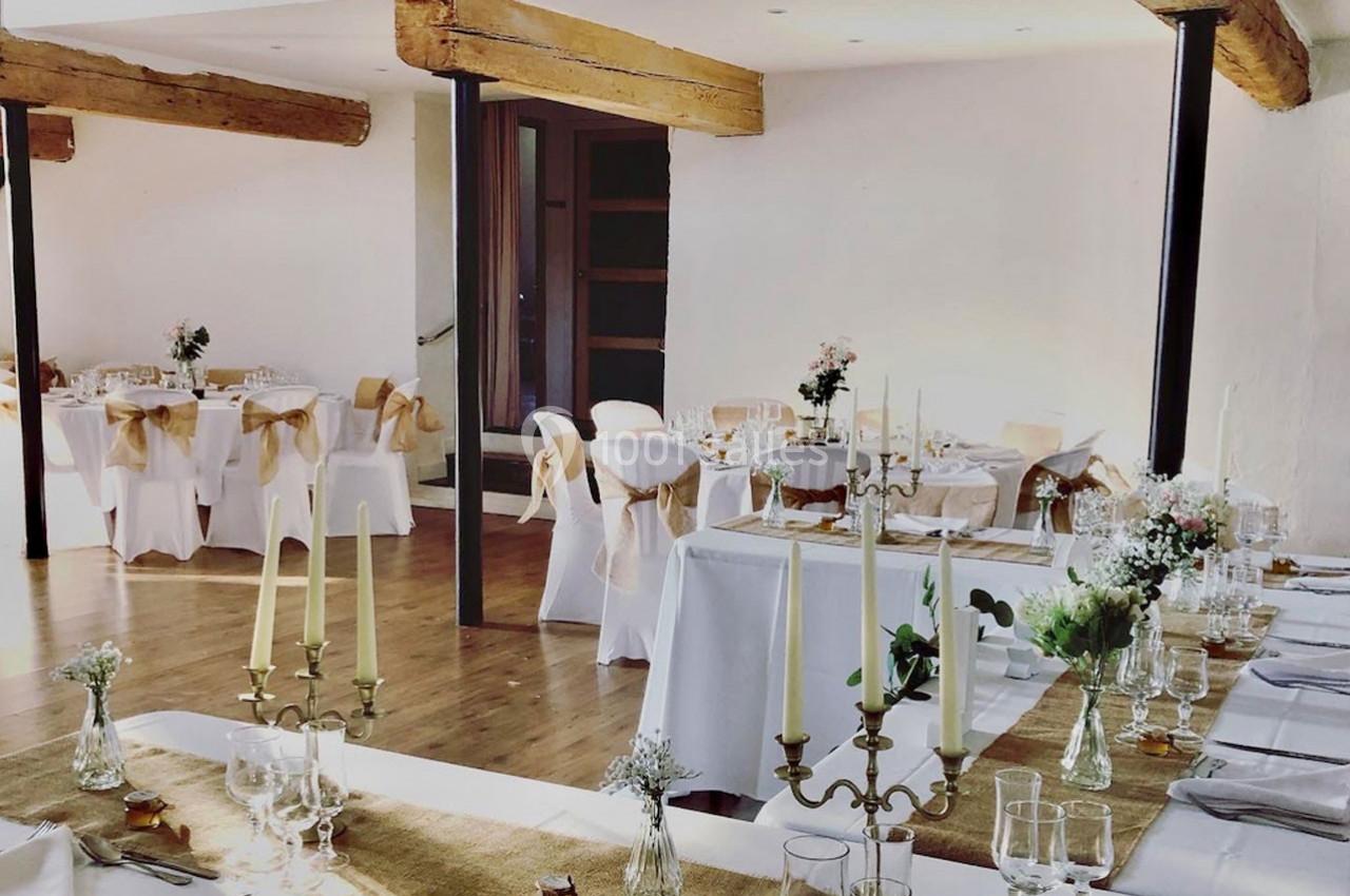 Salle de réception décorée avec des tables dressées, nappes blanches, chandeliers et chaises ornées de rubans beiges.