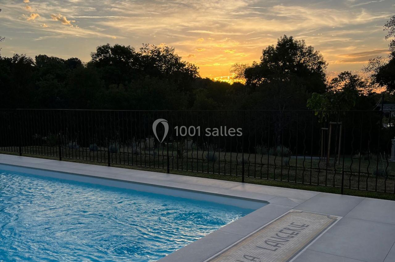 Piscine extérieure avec eau calme au premier plan, entourée de végétation, sous un coucher de soleil.