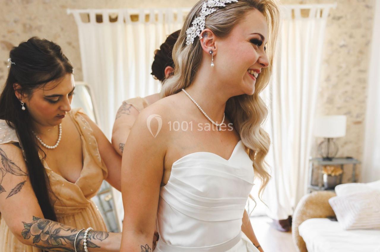 Une mariée souriante en robe blanche est aidée par des demoiselles d'honneur dans une pièce lumineuse.