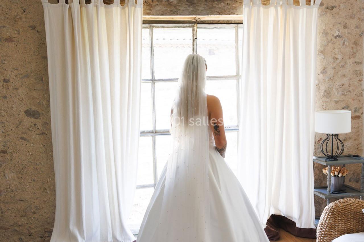 Une mariée en robe blanche et voile regarde par une fenêtre encadrée de rideaux dans une pièce en pierre.