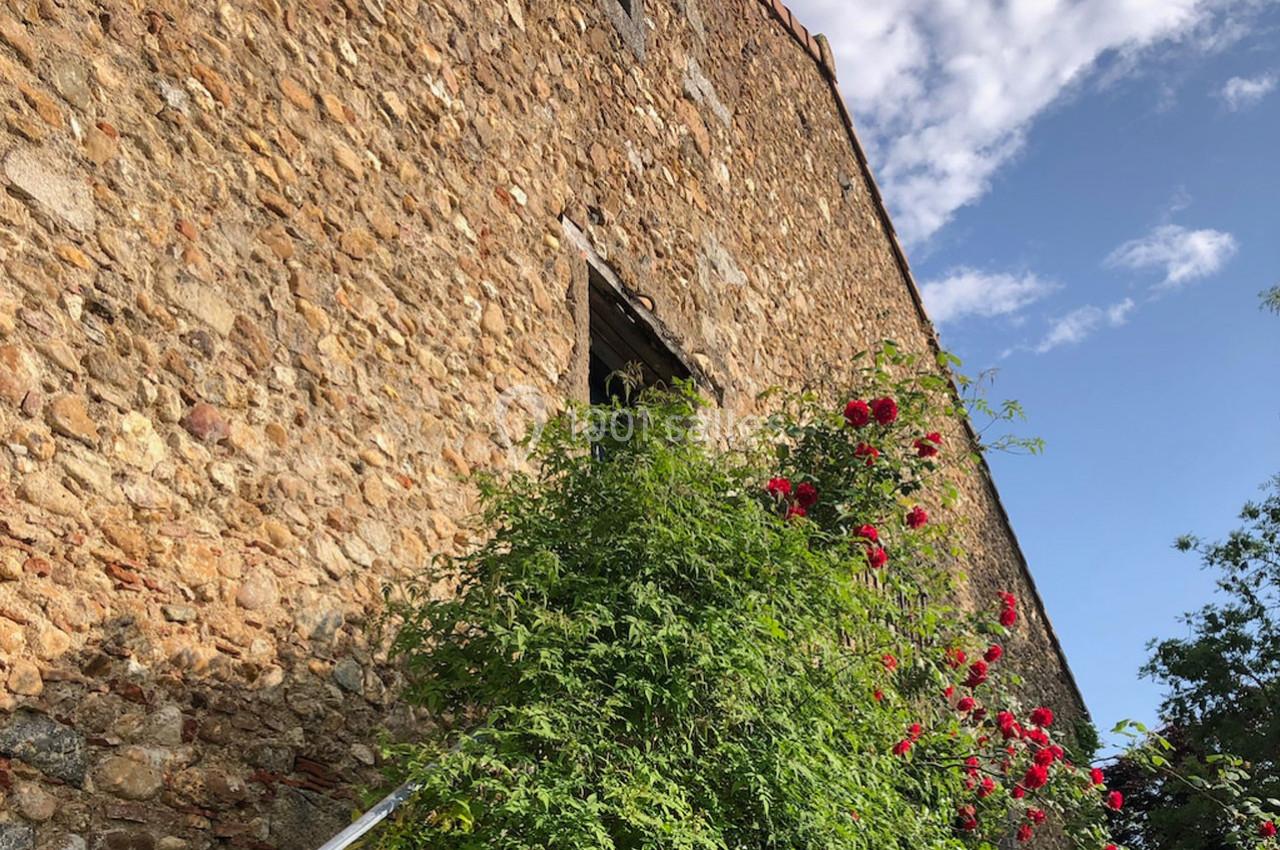 Mur en pierre avec une fenêtre, entouré de rosiers fleuris et d'un ciel partiellement nuageux en arrière-plan.