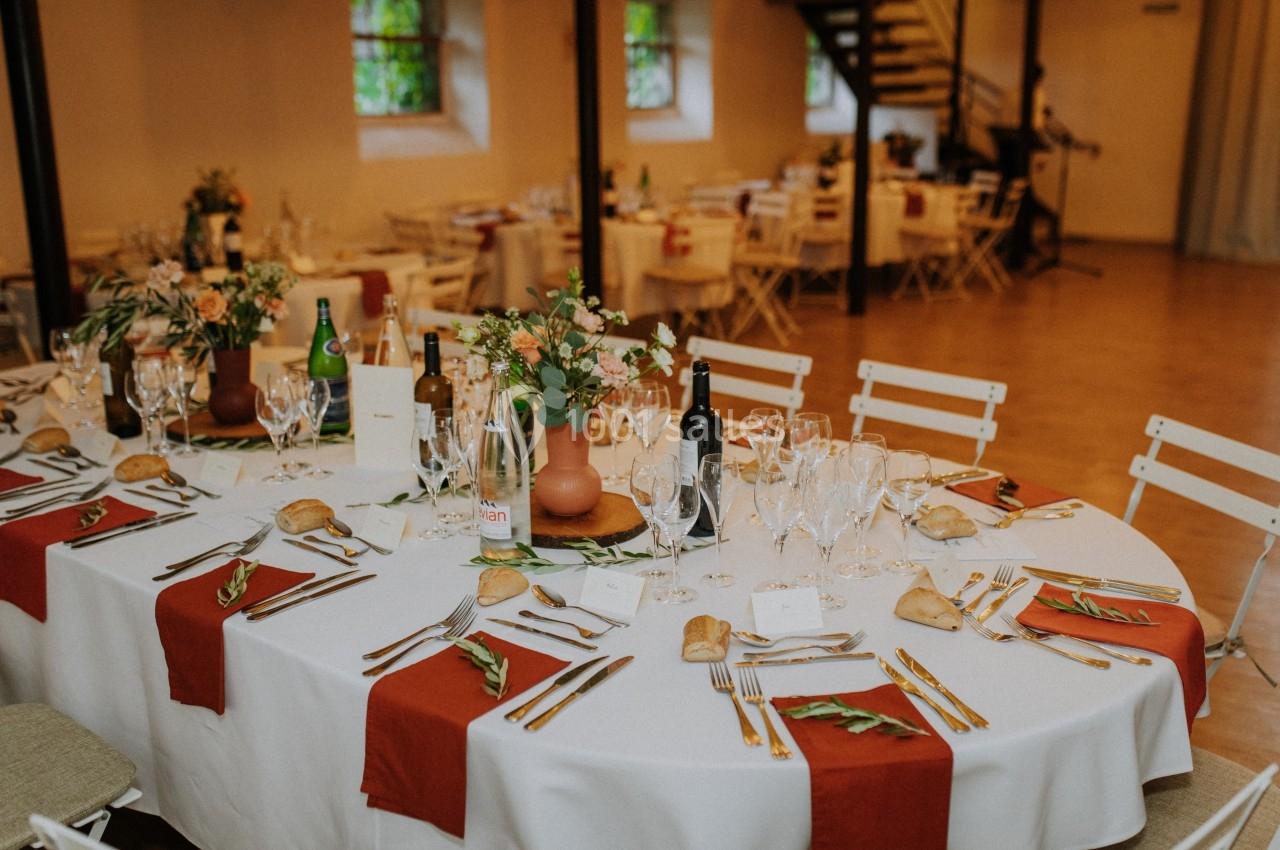 Table ronde décorée pour un repas, avec nappes blanches, chemins de table rouges, vaisselle et arrangements floraux.