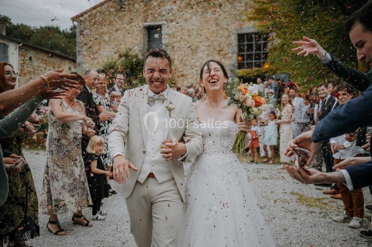 Un couple de mariés souriant marche sous une pluie de confettis, entouré d'invités devant un bâtiment en pierre.