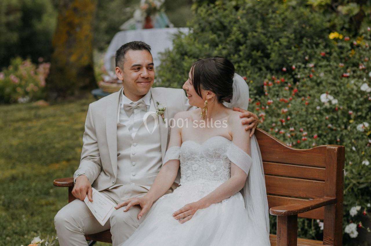 Un couple de mariés assis sur un banc en bois dans un jardin verdoyant, entouré de fleurs.