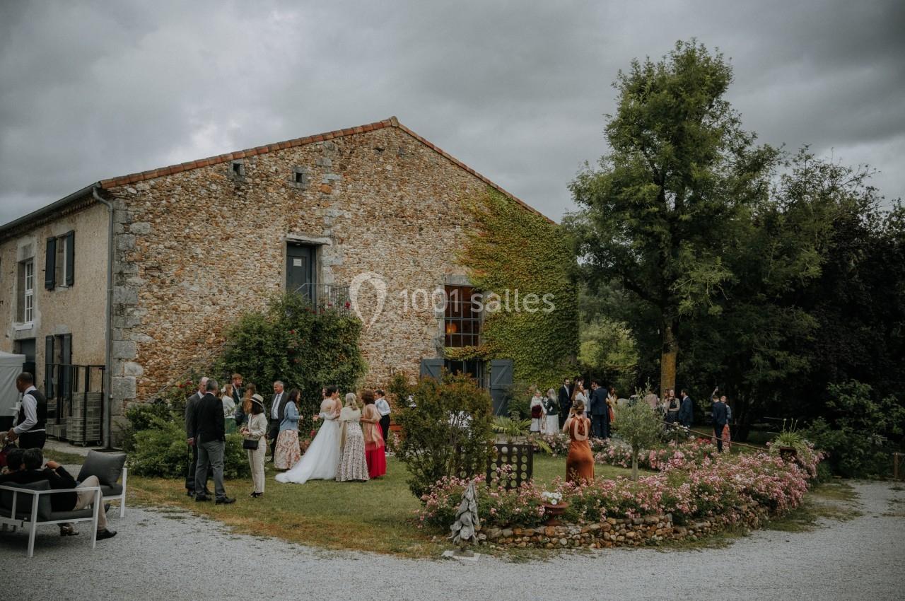 Groupe de personnes rassemblées dans un jardin fleuri devant une maison en pierre sous un ciel nuageux.