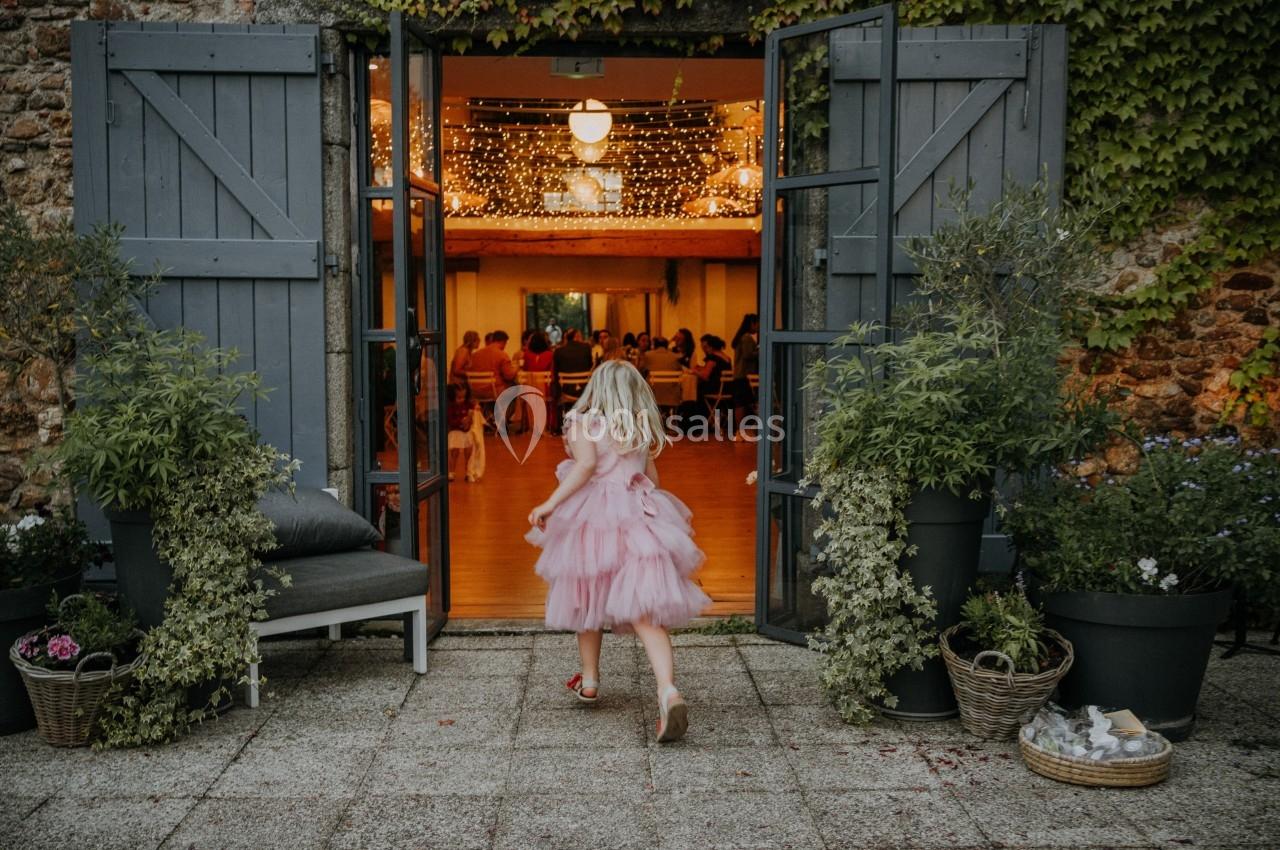 Une fillette en robe rose entre dans une salle éclairée, vue depuis une terrasse ornée de plantes en pots.